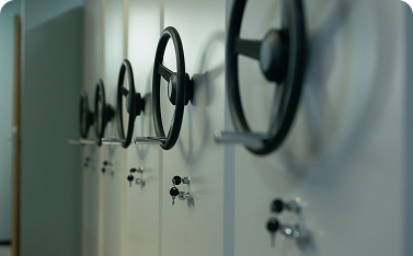 A row of large, black circular handles (wheels) and key locks extending from the side of a set of white, compact shelving units, suggesting secure document storage.