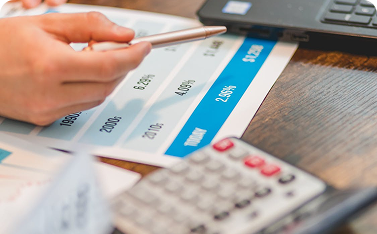 Close-up of a hand holding a pen, pointing at a printed financial chart that includes percentages and the word "TODAY." A calculator rests on the table in the foreground.