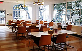 Bright, modern cafeteria or dining hall with wooden floors, tall windows, and multiple square tables paired with brown and orange chairs.